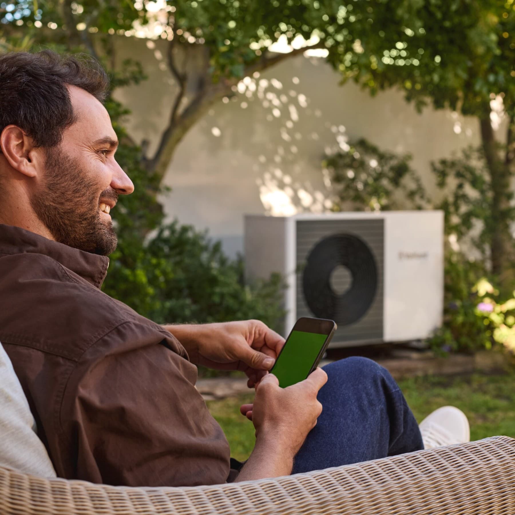 Mann sitzt mit Handy im Garten neben einer Vaillant Wärmepumpe