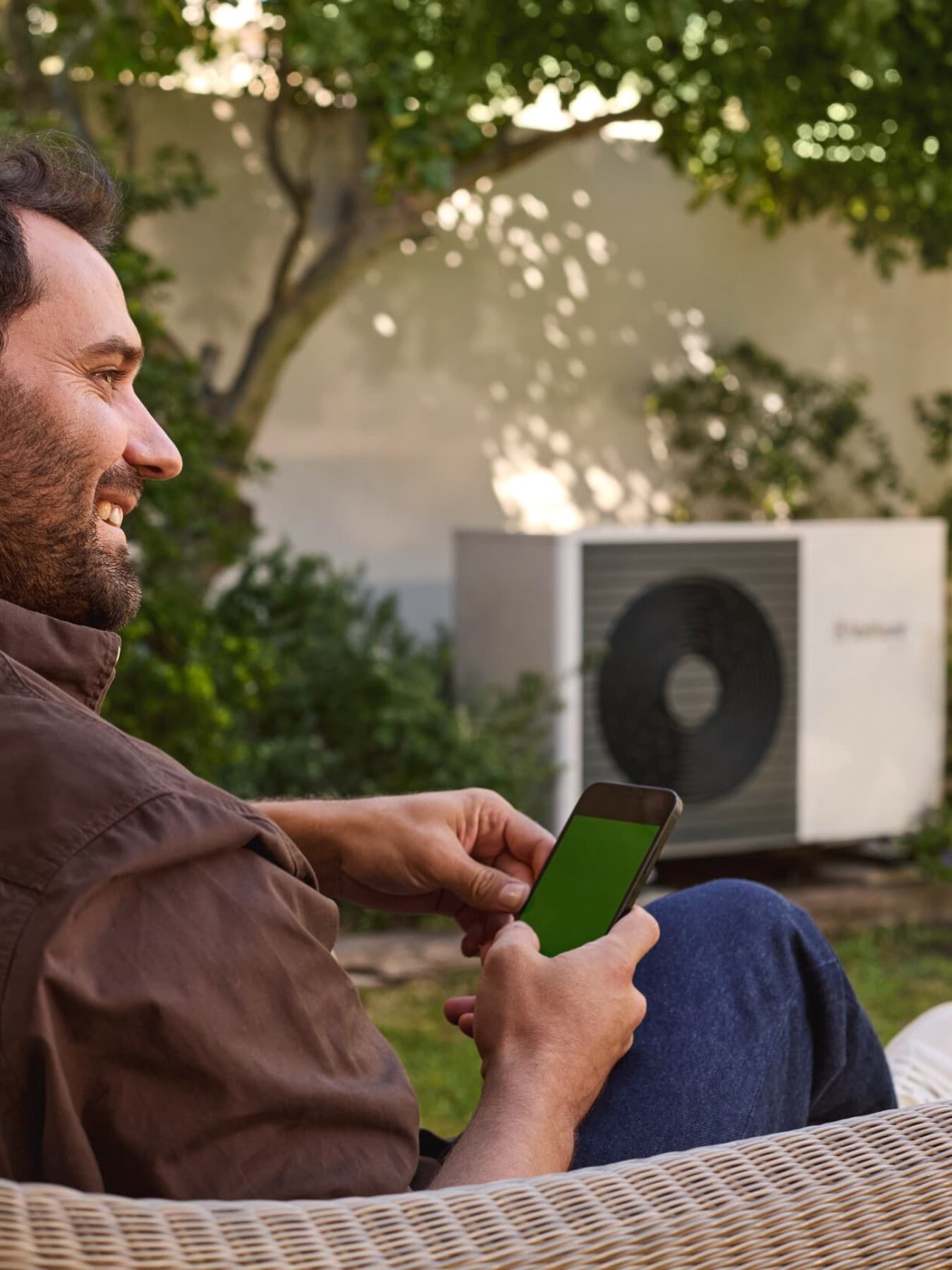 Mann am Handy sitzt im Garten vor einer Wärmepumpe