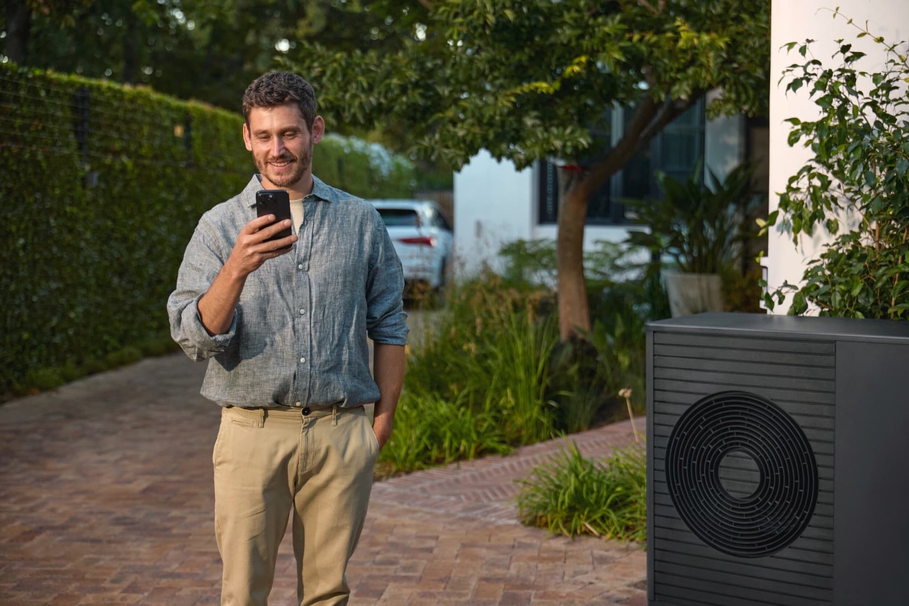 Mann, der neben einer Wärmepumpe steht und auf ein Smartphone schaut