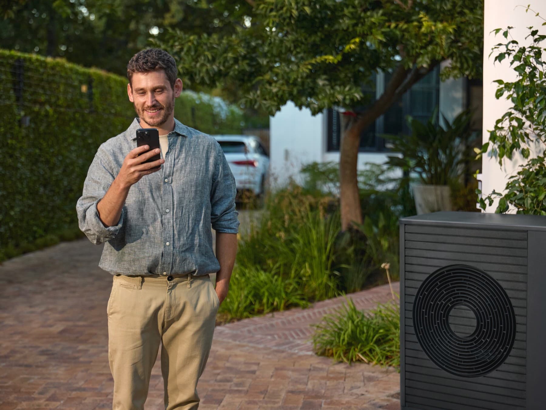 Mann, der neben einer Wärmepumpe steht und auf ein Smartphone schaut