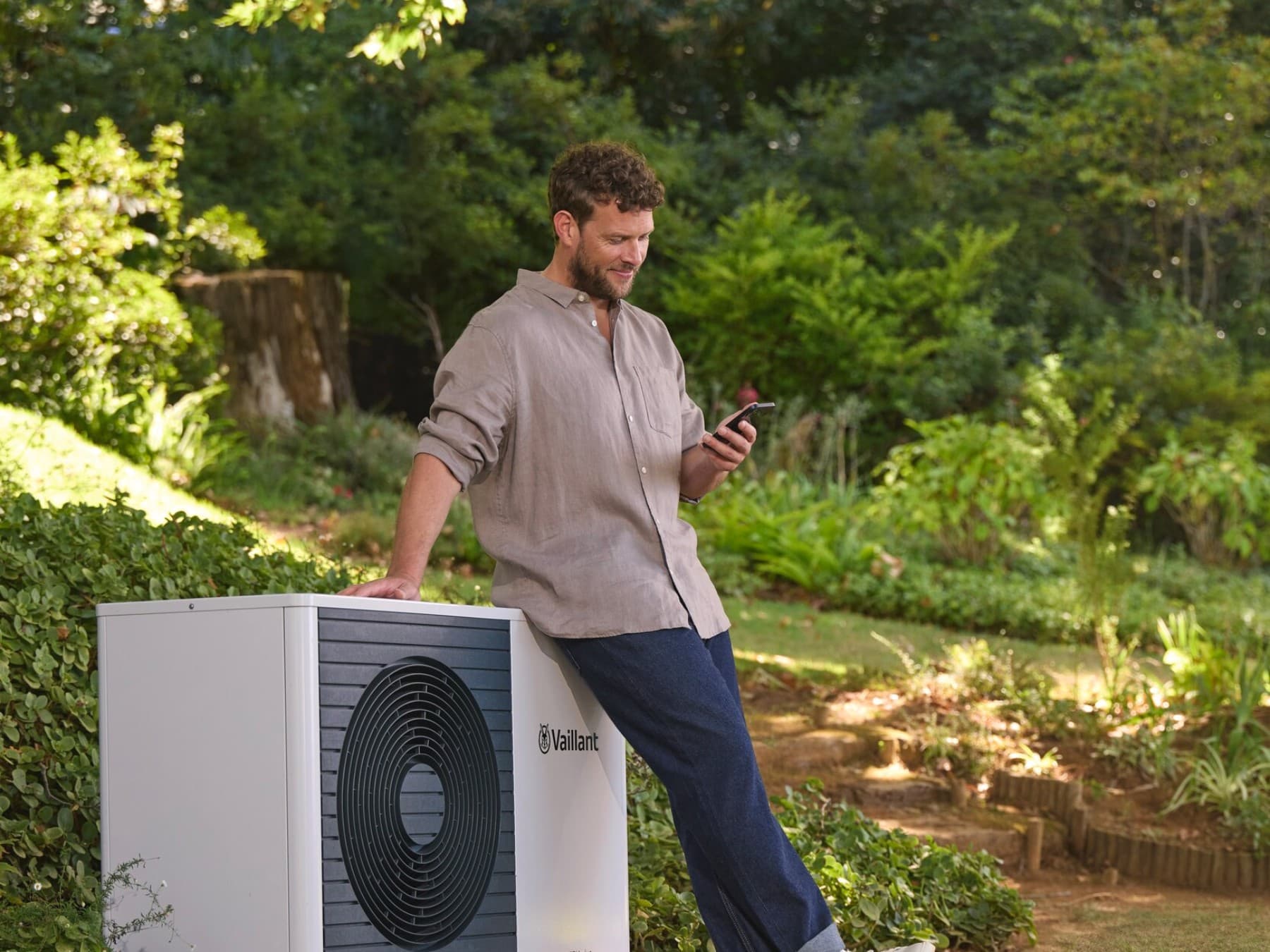 Mann schaut auf sein Smartphone und steht neben der Wärmepumpe im grünen Garten