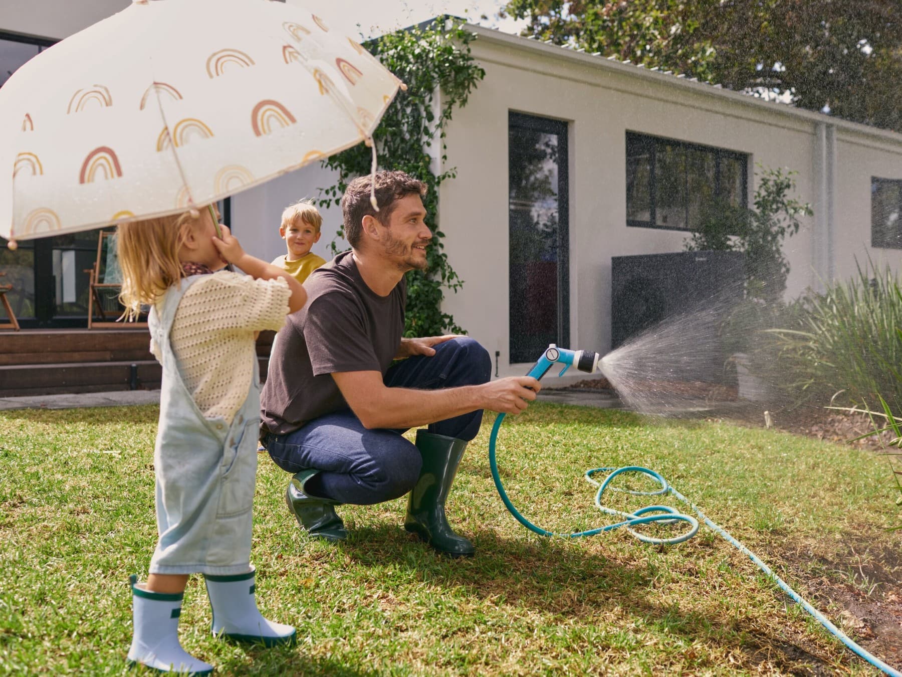 Vater gießt zusammen mit den Kindern die Blumen im Garten vor einer Wärmepumpe