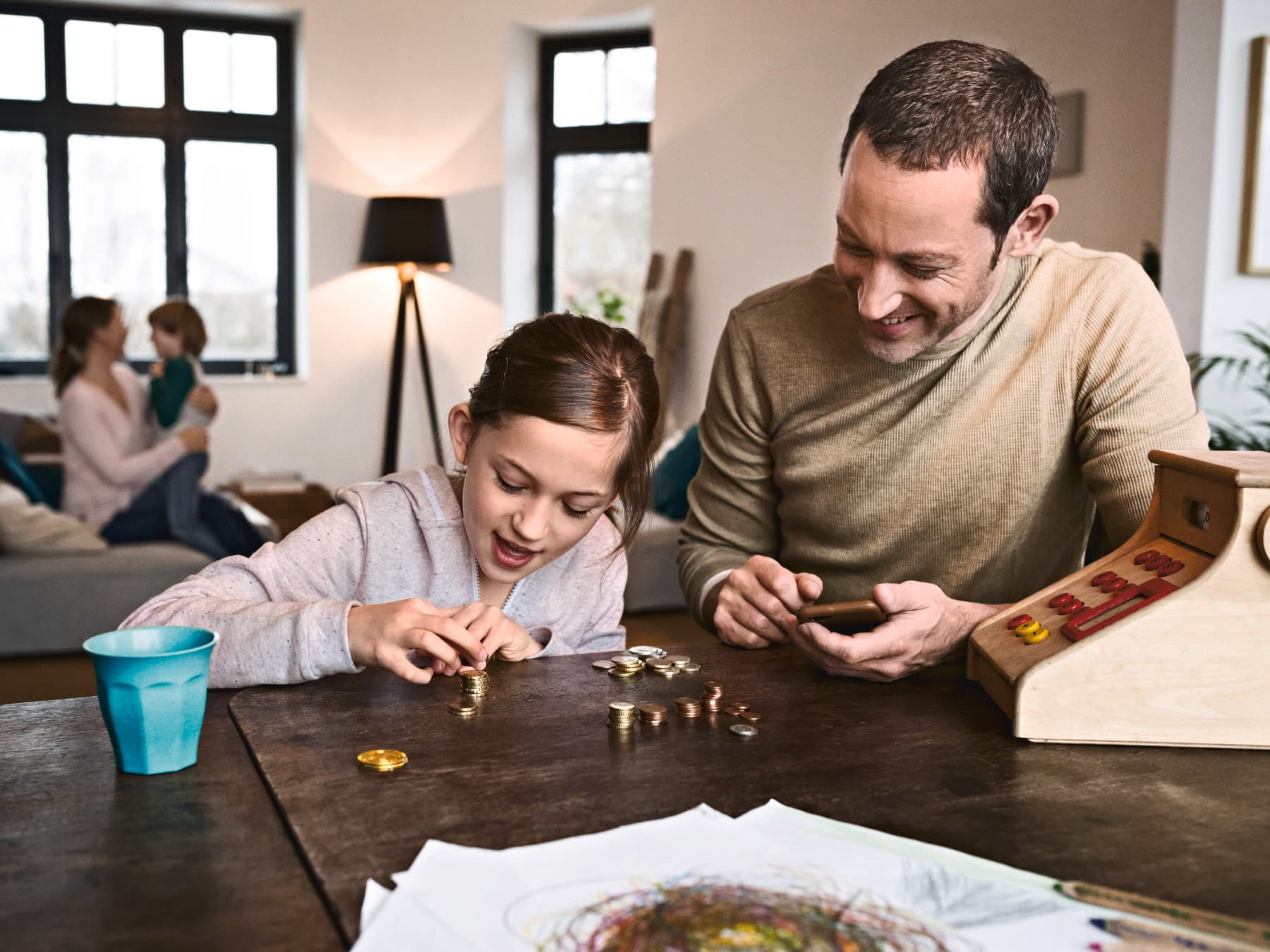 Mädchen stapelt Geldmünzen auf einem Tisch, neben ihr sitzt der Papa und schaut aufs Handy, im Hintergrund toben Mama und Bruder auf der Couch.