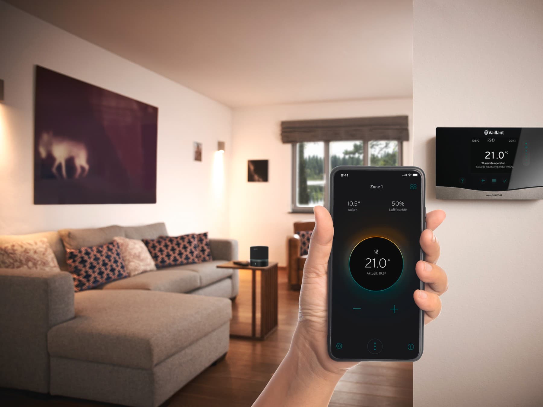 Eine Hand vor einem Vaillant Regler, die ein Smartphone hält, auf dem die myVaillant App zu sehen ist